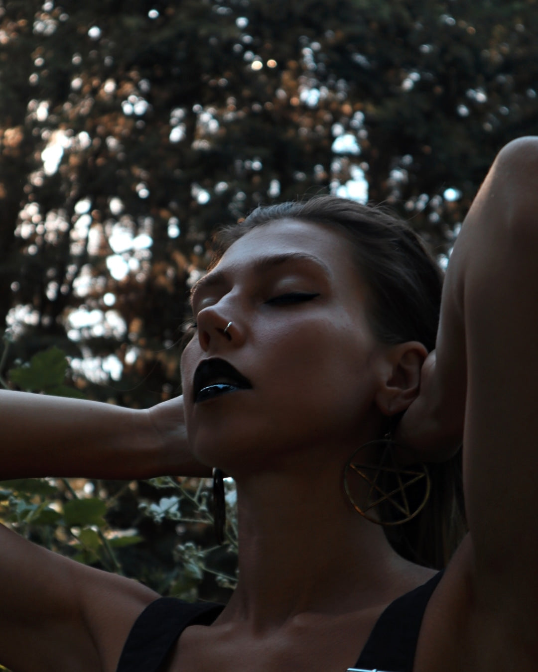 Woman with dark makeup and a pentagram earring against a blurred natural background