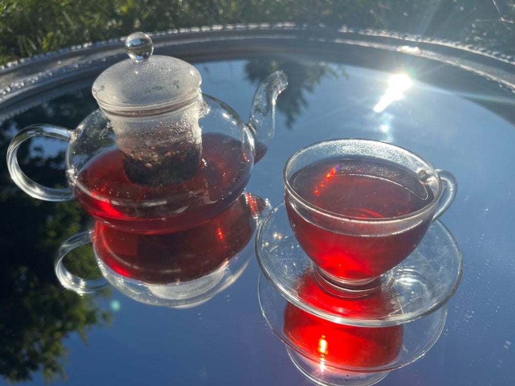 INNERGLO TEA Herbal Blend A summery lifestyle photo with a teapot and cup filled with radiant pink INNERGLOW TEA, highlighting its rich color and fruity-spiced aroma.